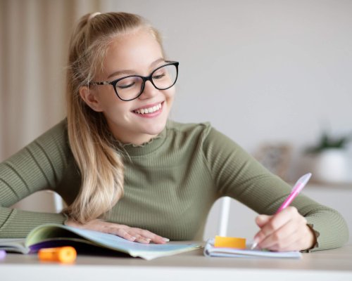 Exciting school education concept. Cute blonde school girl in glasses doing homework and smiling, sitting at table with book, writing essay or making test, home interior, copy space