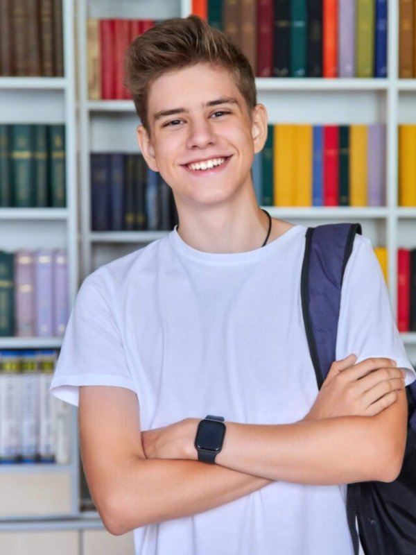 Single portrait of smiling confident male student teenager with backpack with crossed arms looking at camera in the library. School, education, knowledge, adolescence concept